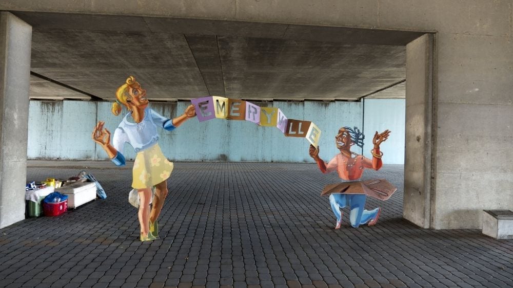 A colorful metal statue of two people holding a sign that says “Emeryville.”