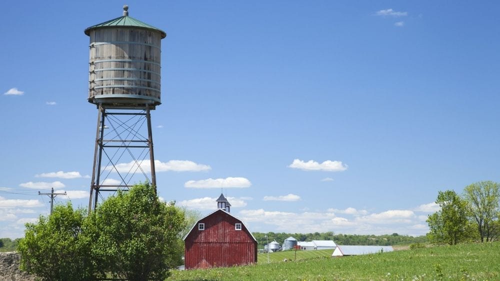 An old water cistern sits next to a red barn in a rural area.