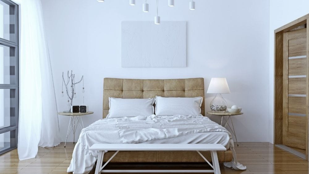 A bed with white sheets and a wooden headboard. The floor beneath is laminate.