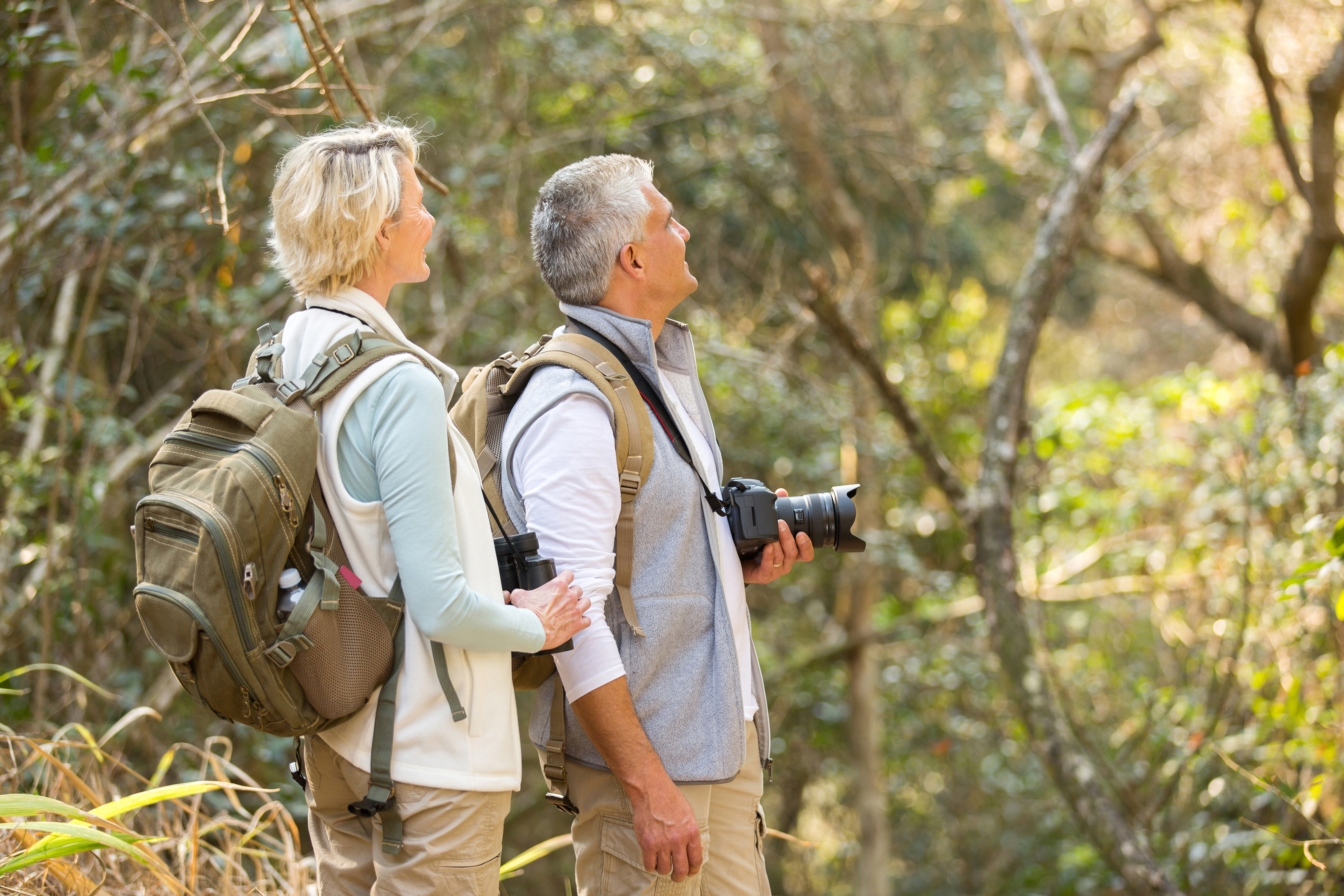 bird watching seniors with backpack, camera, and binoculars in forested area 