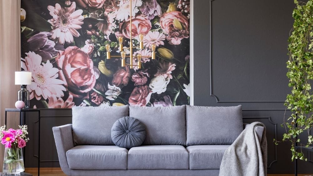 Pink floral, dramatic wallpaper with a dark background, next to a dark wall with trim. Grey sofa in the foreground, with a throw pillow and blanket