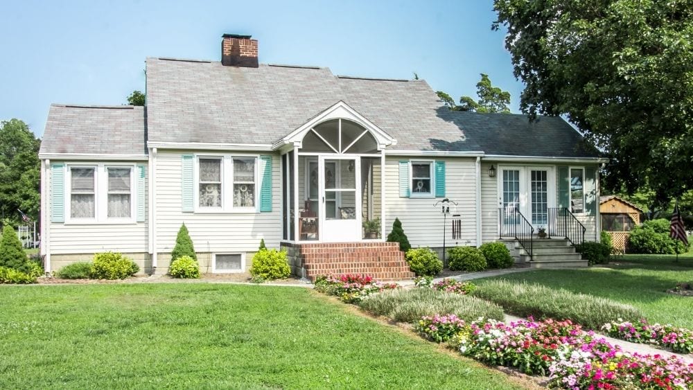 A colonial style house with a grey shingled roof and light blue siding. A brick path lined with colorful flowers leads to the door.