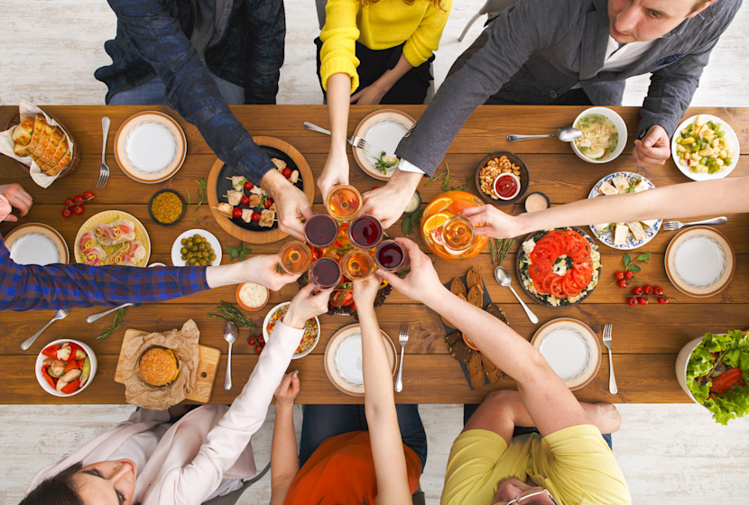 A group of people holding glasses for a toast, over a wooden dining table, laden with different dishes of food.