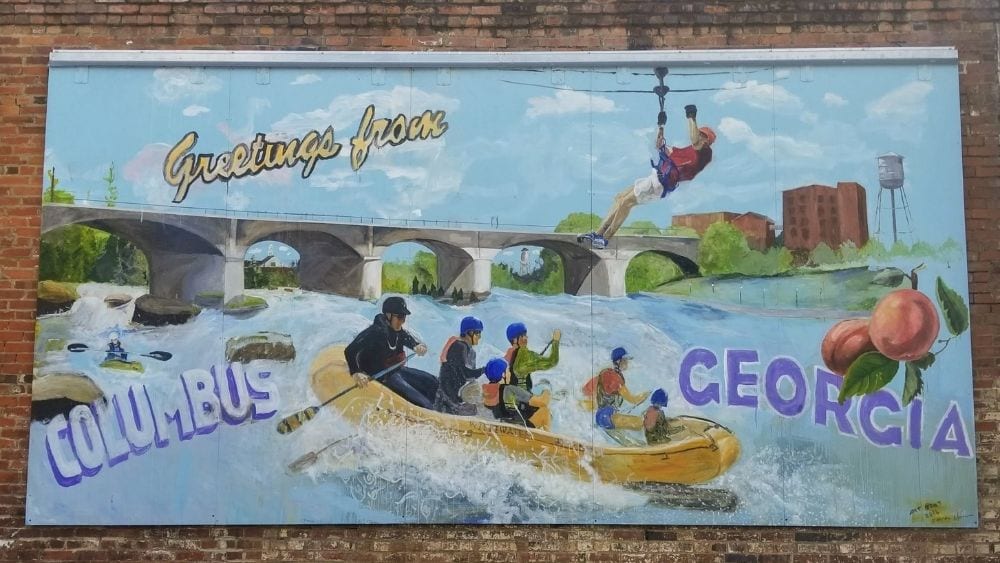 A mural on a brick wall depicts people white water rafting. The mural reads, “Greetings from Columbus, Ohio.”