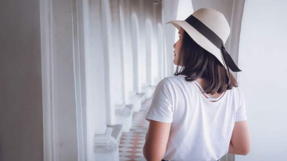 Woman wearing a white hat with a black bow, and a white shirt, walking down a hallway or white archway windows.