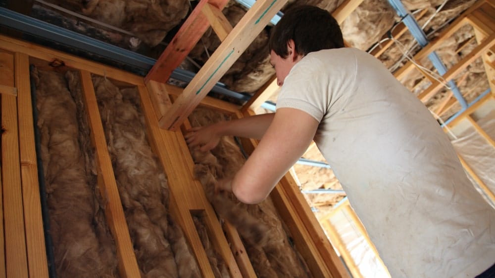 A young man puts insulation batting in between a wall’s wood frame in a new construction home