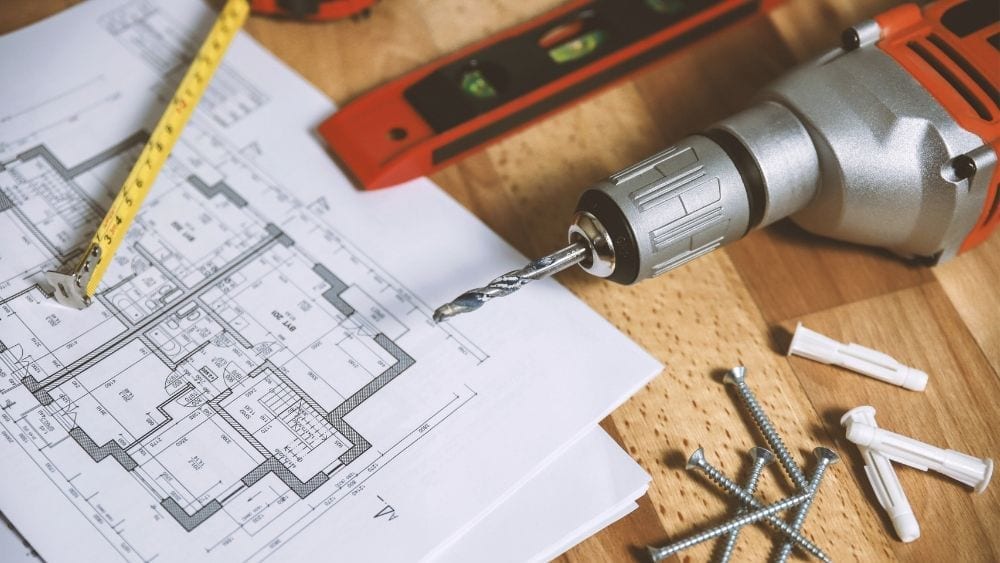 House plans on a table with a drill, drill bits, a measuring tape and a level lying next to it.