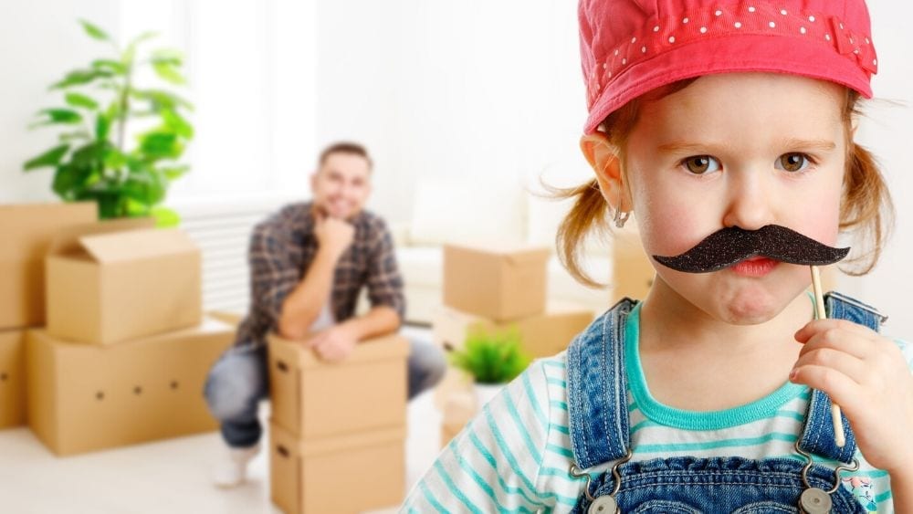 Child with paper moustache in foreground with dad with boxes in the background