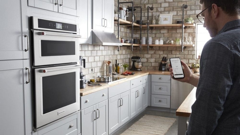A man stands in his kitchen, holding his smartphone and looking at an app that can connect to his kitchen appliances.