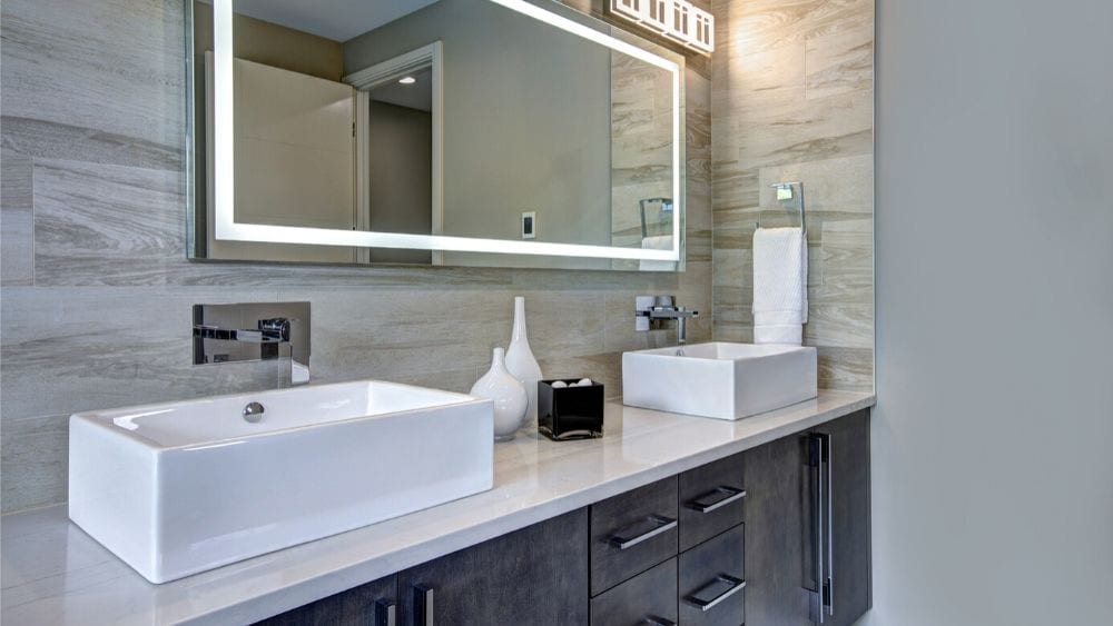 dual sinks on a bathroom vanity with dark cabinets and quartz counters. There is a long, rectangular mirror with built-in lighting on the edge.
