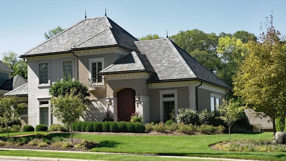 A large house made of brick and stucco with a grey slate roof, with manicured lawns, trees and hedges