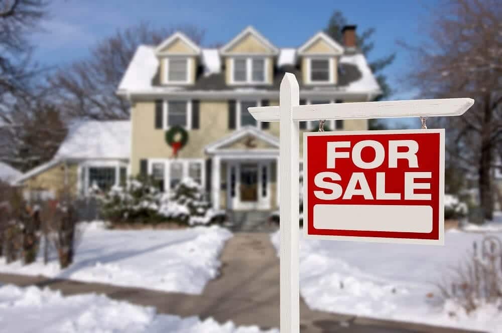 Snowy home decorated for the holidays with a red “For Sale” sign in the foreground