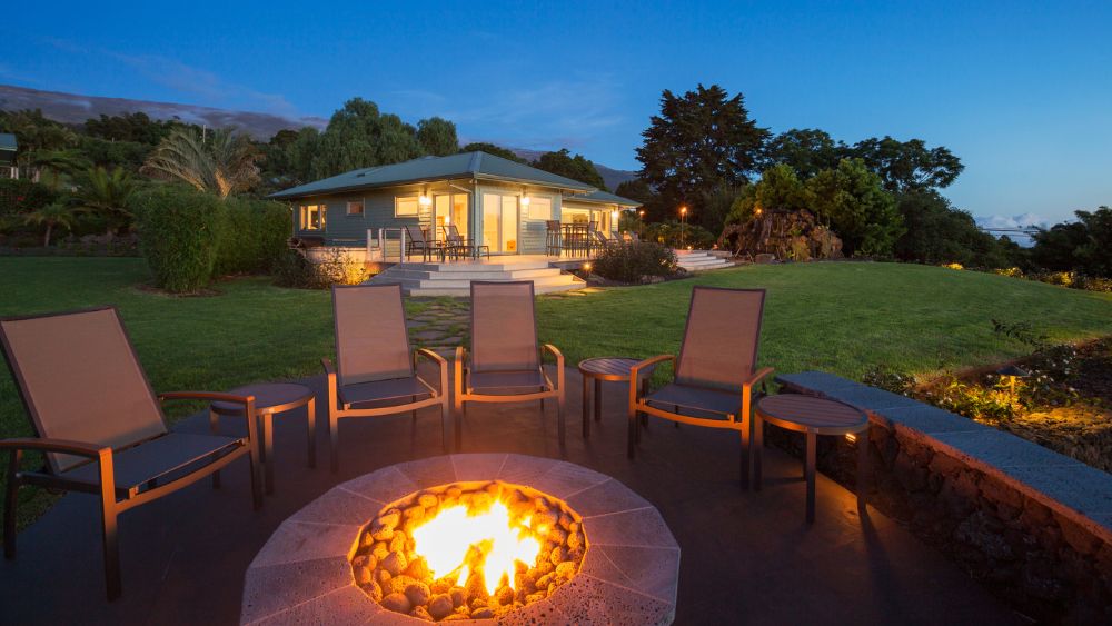 A lit backyard fire pit at dusk surround by chairs, with a large yard and house in the background.