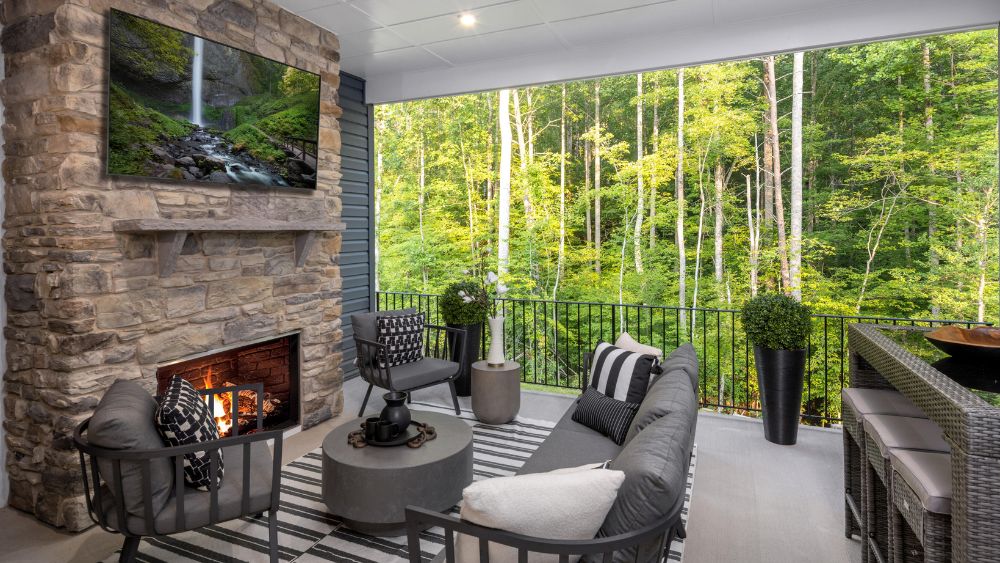 A patio looking out into a forest with a sitting area, a stone fireplace, and a flat screen tv mounted.