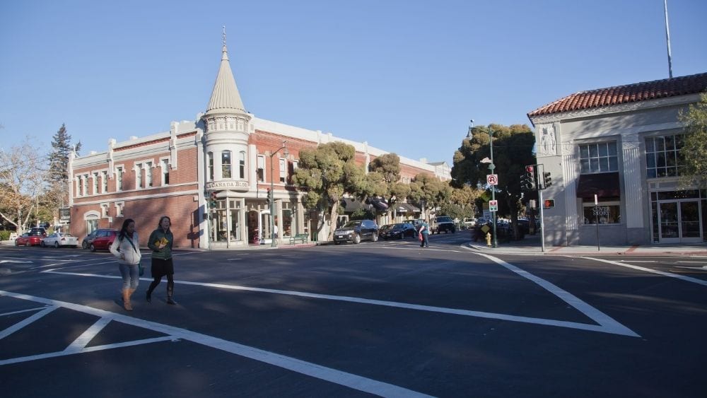 Street intersection with people walking. A stately building is cattycorner from the viewer and looms above the other buildings.