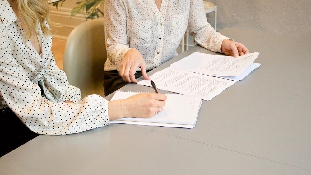 Two people sitting at desk talking over documents.