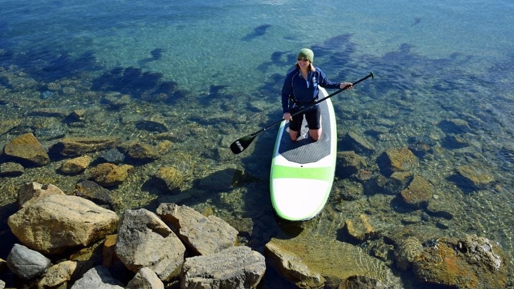 A woman kneeling on a paddle board in shallow water.