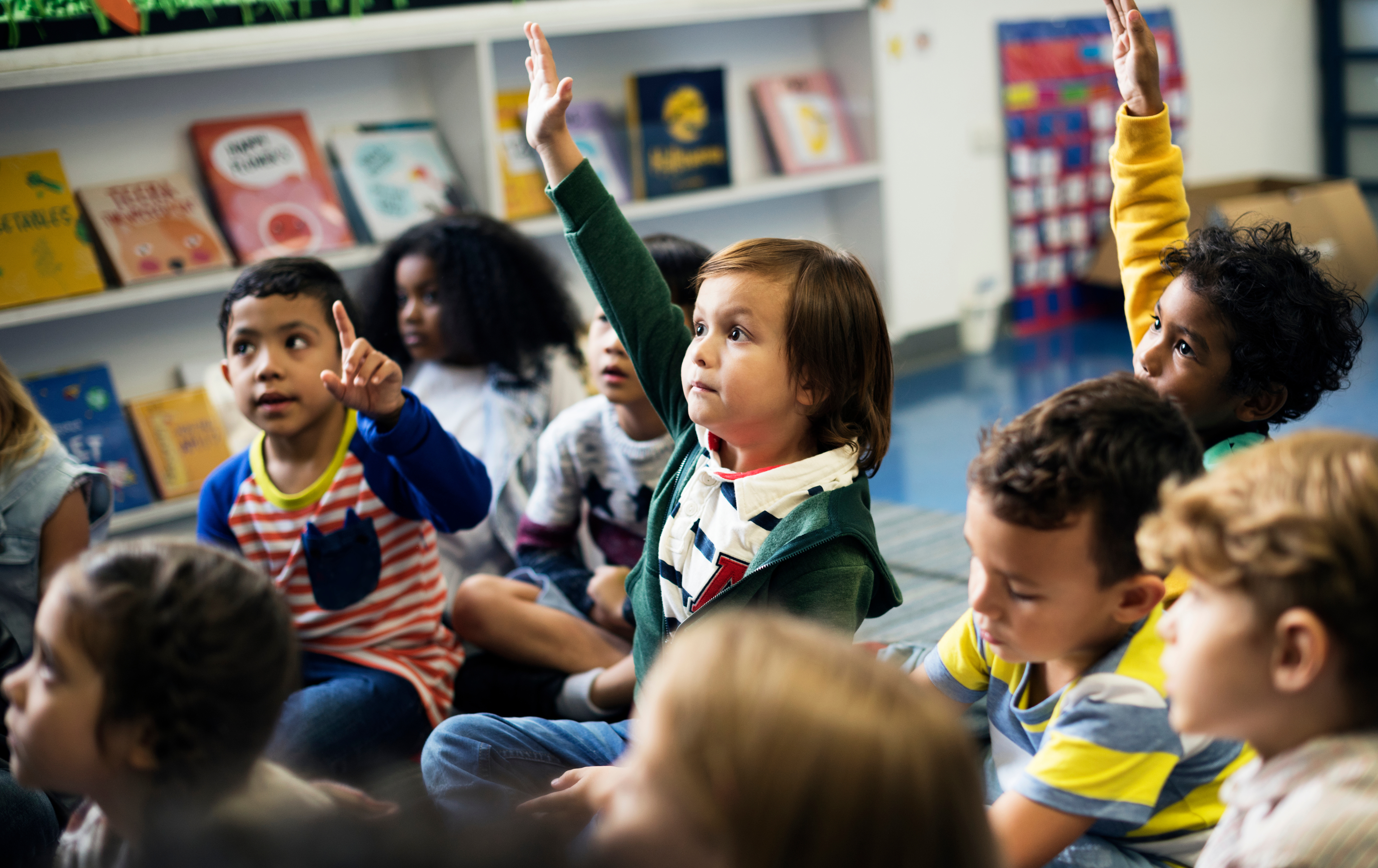 Young children sitting on the floor in a school setting; one child has his hand raised