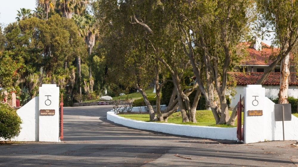 Two white stucco markers with red clay tops indicate the entrance to a paved road lined with trees covered in Spanish moss.