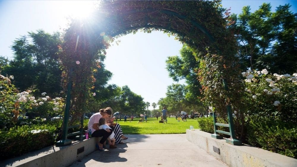 A shrub archway with people sitting beneath, leading into an open park.