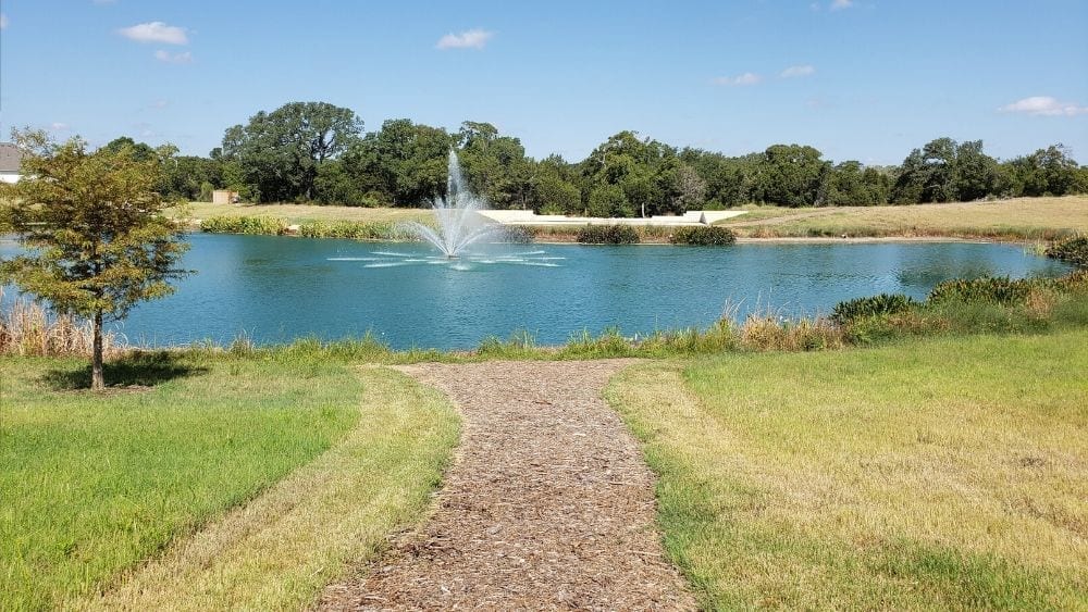 A gravel path leading up to a large fishing pond with a fountain in the middle.
