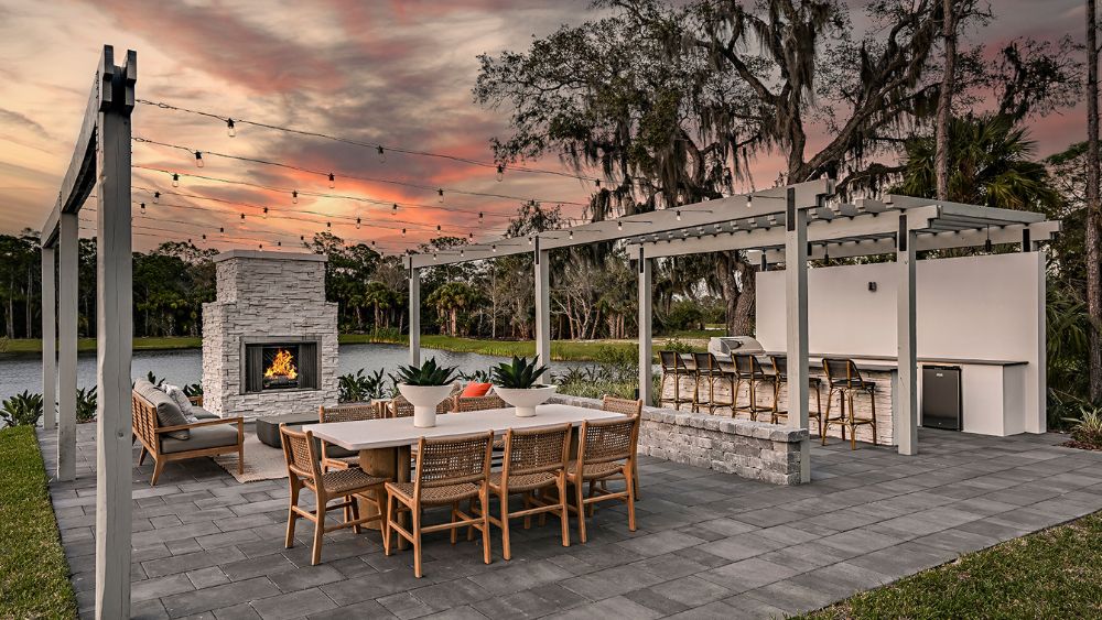 A backyard at sunset with an outdoor fireplace, table, bar, and pergola.