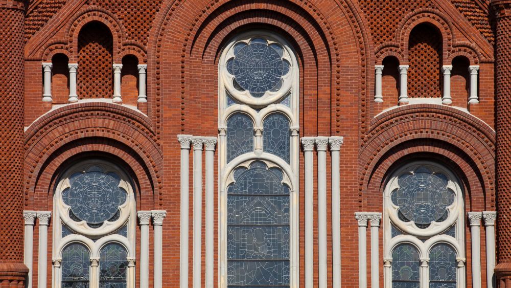 Church windows in arches, with two shorter ones on the outside and a tall arch in the middle, with red brick facade.