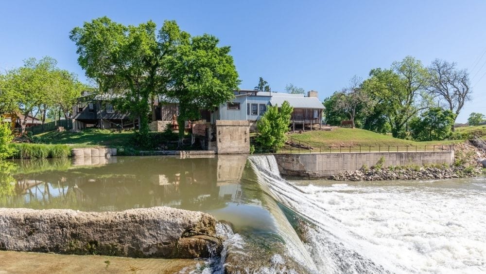 A dam  with water rushing over it, and a metal tin building in the background.