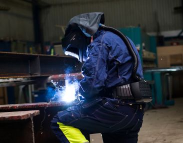 Welder in blue and yellow protective clothing wearing a 3M Adflo PAPR connected to a 3M Speedglas helmet. He's kneeling and performs a weld on a metal beam in a dark workshop.