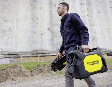 Worker in navy blue and grey workwear carrying an ESAB portable welder outdoors near a concrete wall.