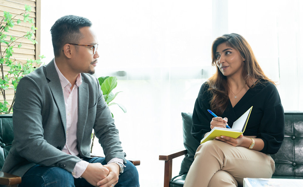 Two colleagues sat in leather chairs, facing each other and discussing a business case for coaching,