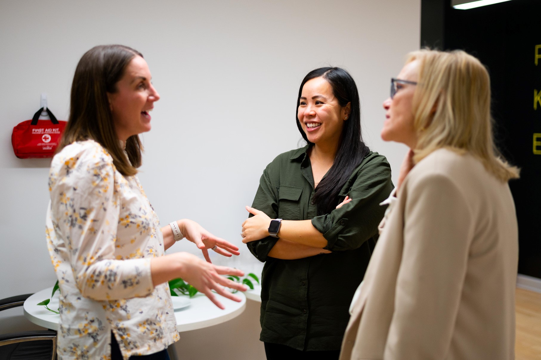 Three women stood talking in an office space.