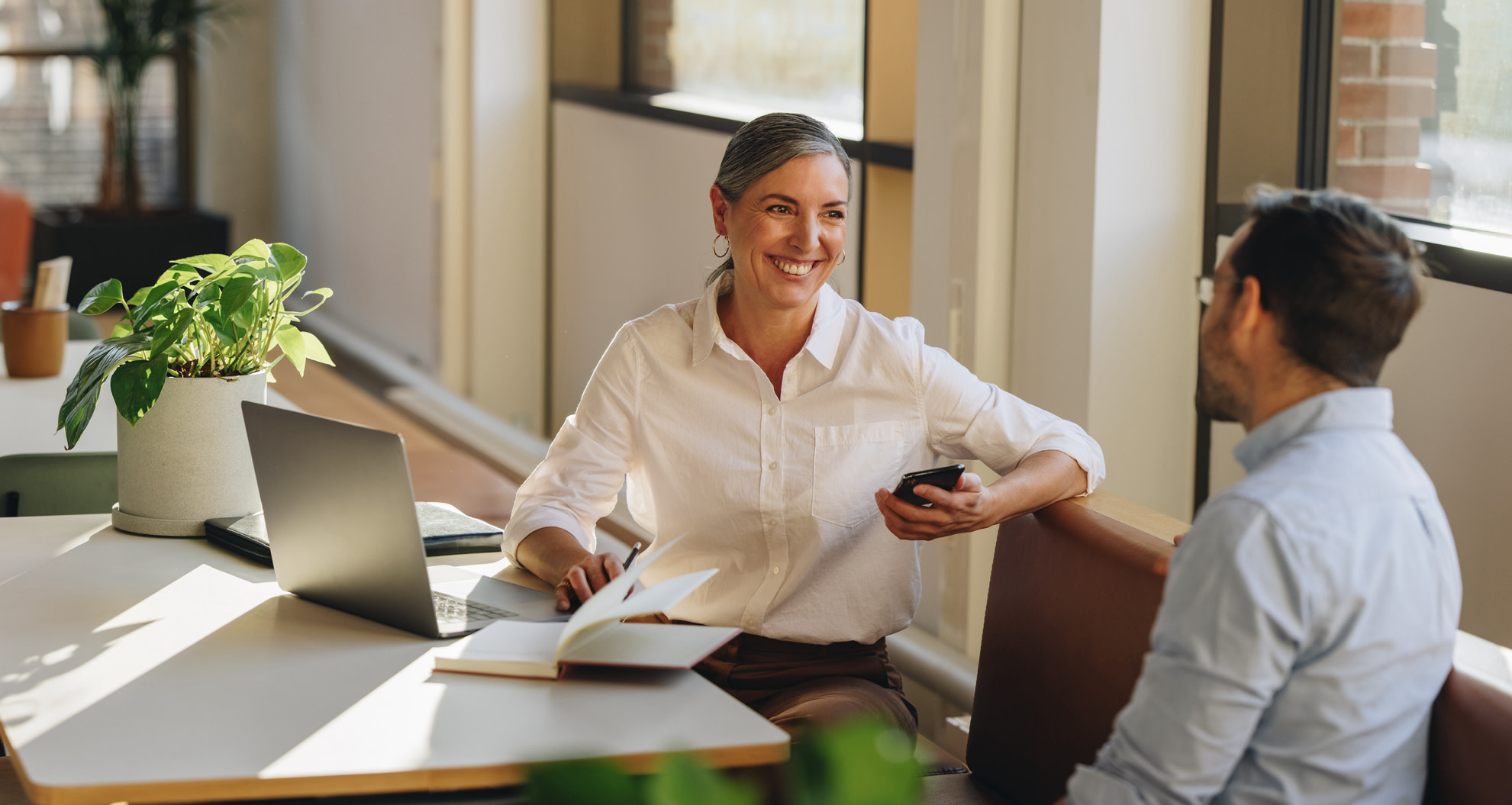 A woman in a white top talking to a man in a light blue shirt, both sitting at a desk with a plant and a laptop.