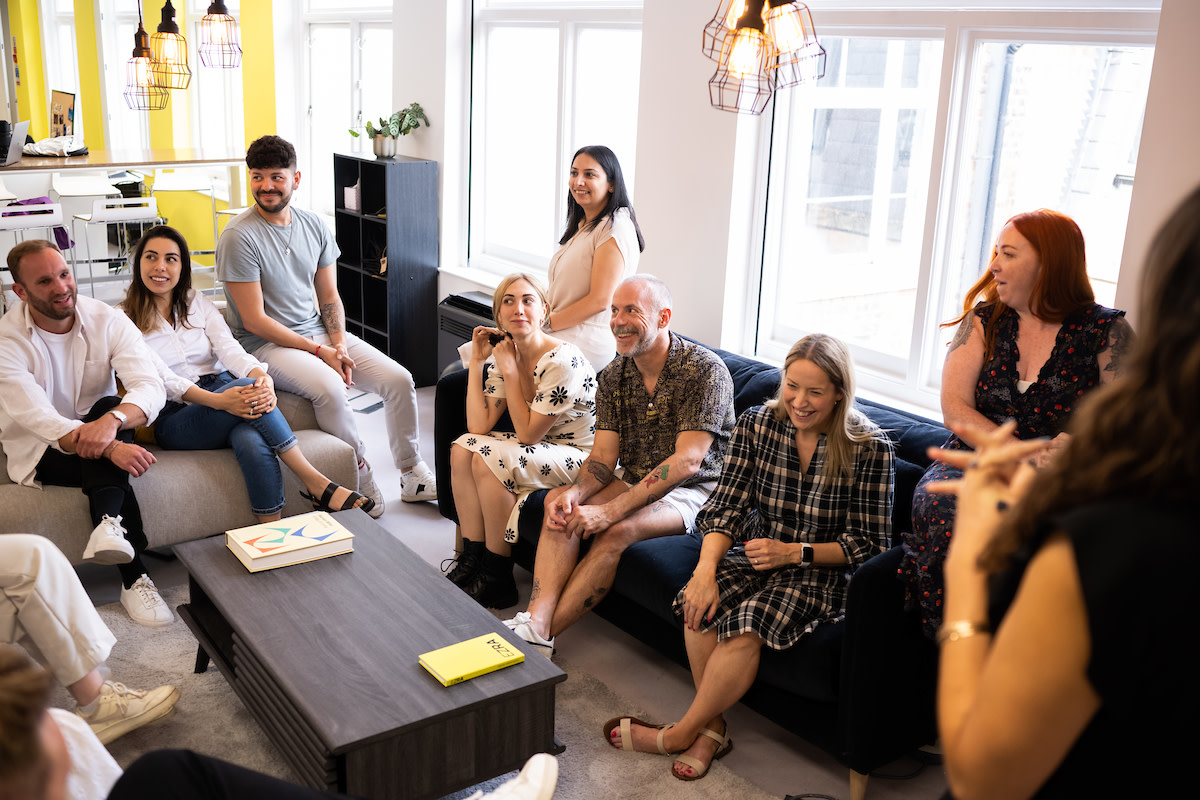 A room full of colleagues gathered around two sofas and a coffee table, discussing organizational performance.