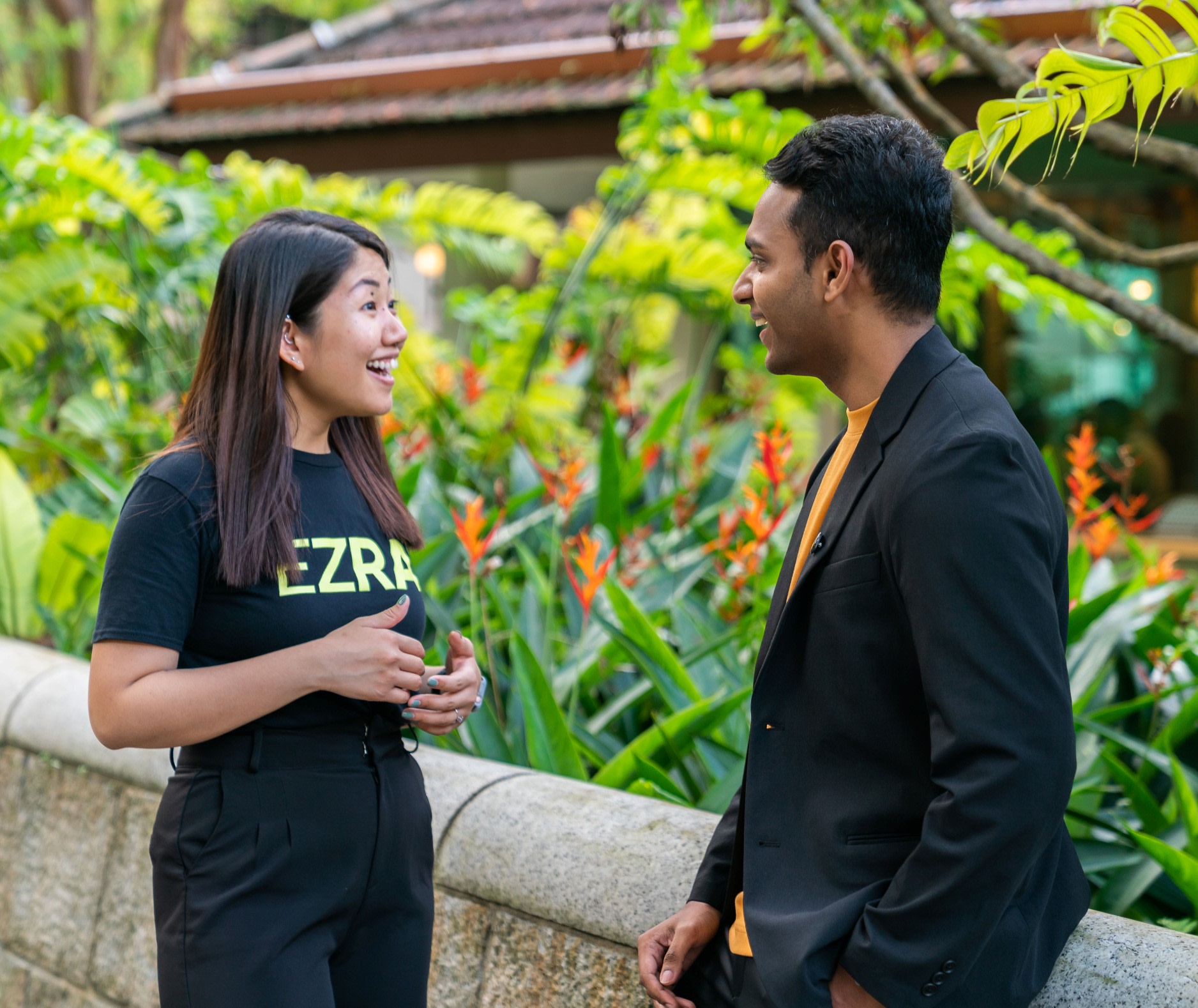 Smiling woman wearing a black EZRA t-shirt, talking to a man wearing a dark jacket over an orange tee.