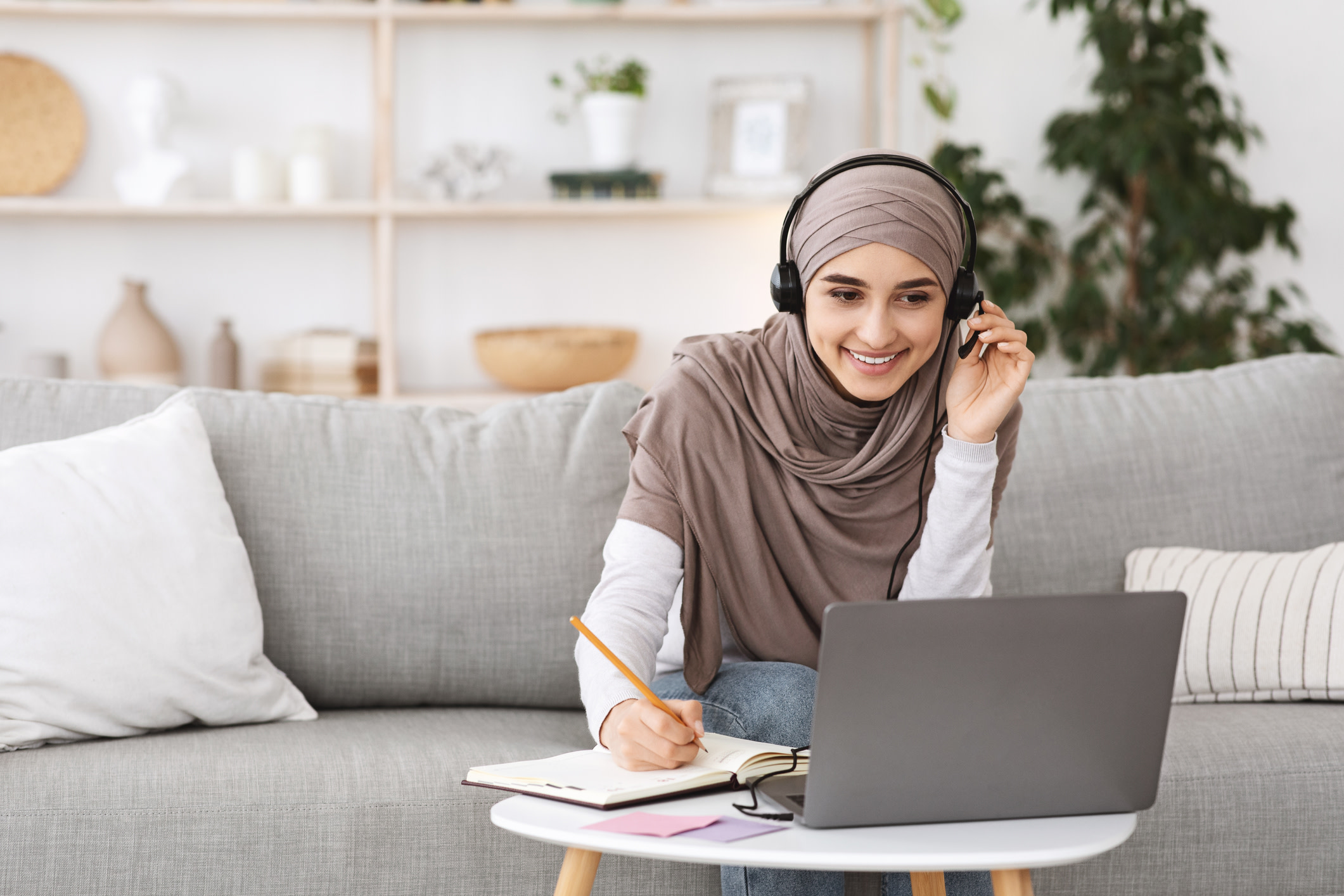 Online Education For Muslim Women. Happy Arabic Girl In Headscarf And Headset Studying With Laptop At Home, Taking Notes While Watching Webinar