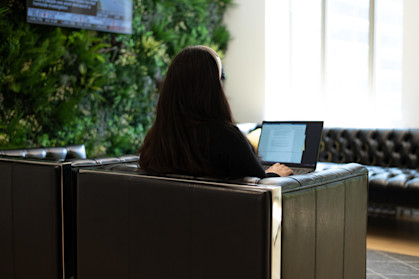 A woman, sat with her back to the camera, in a leather chair working on a word document on her laptop next to a wall of plants and a large screen.