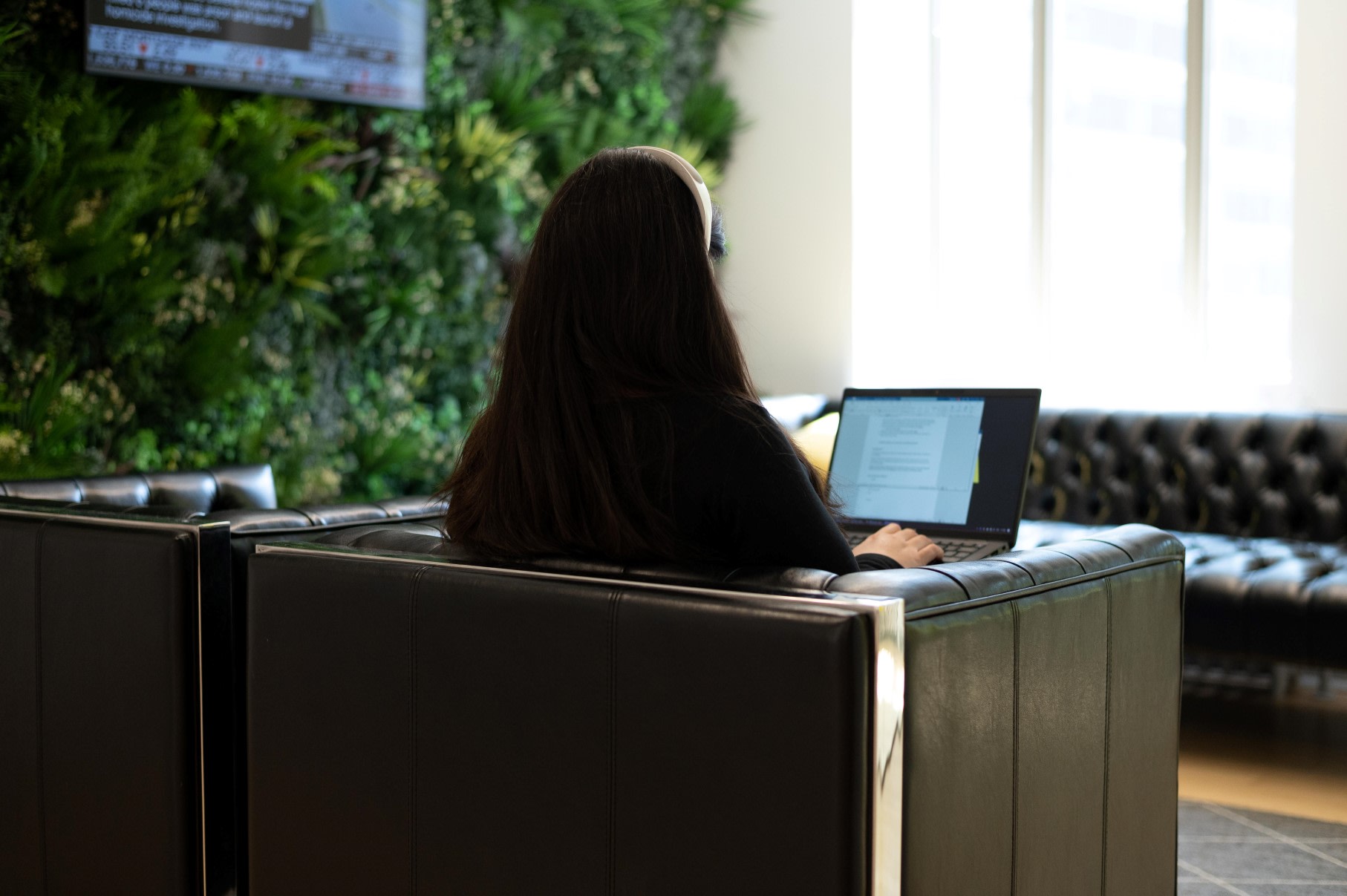 A woman, sat with her back to the camera, in a leather chair working on a word document on her laptop next to a wall of plants and a large screen.
