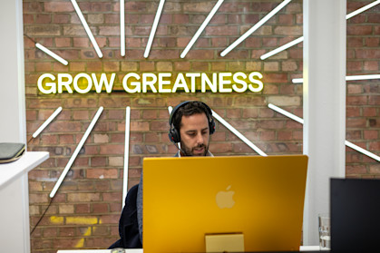 Man sat at his computer in an office with his headphones on, in front of a wall sign that says 'grow greatness'.