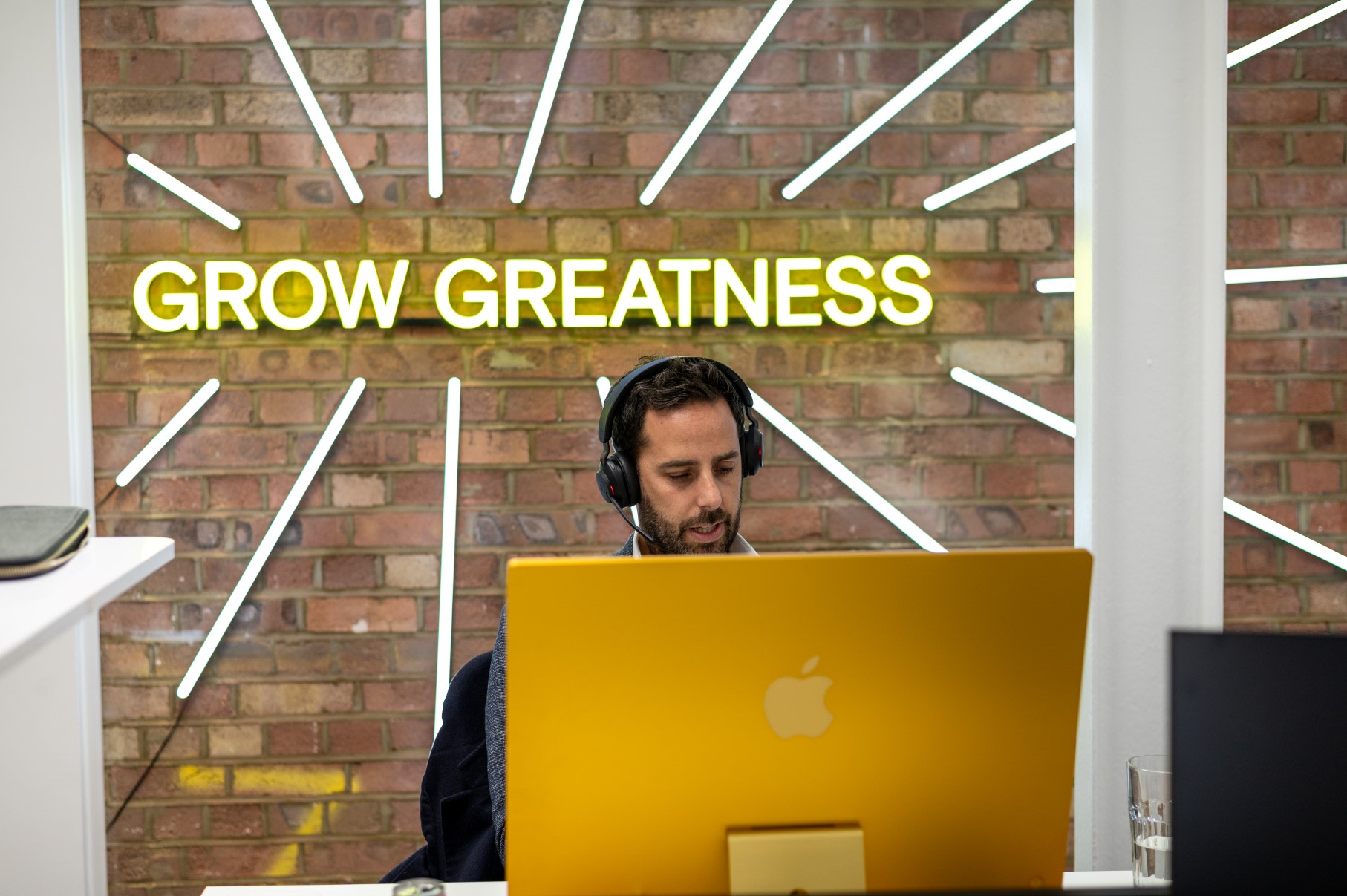 Man sat at his computer in an office with his headphones on, in front of a wall sign that says 'grow greatness'.