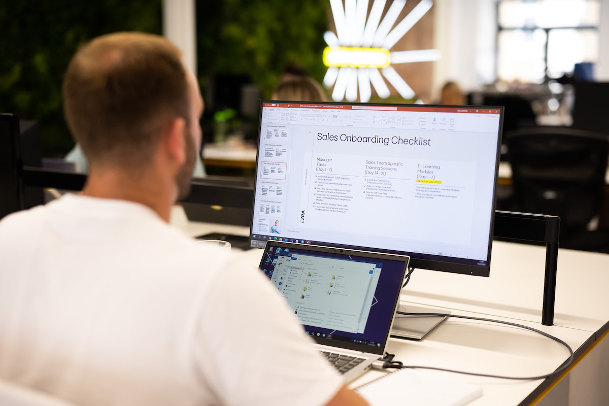 A man sat at his desk, facing his laptop and computer screen that has a presentation slide named 'sales onboarding checklist' visible.