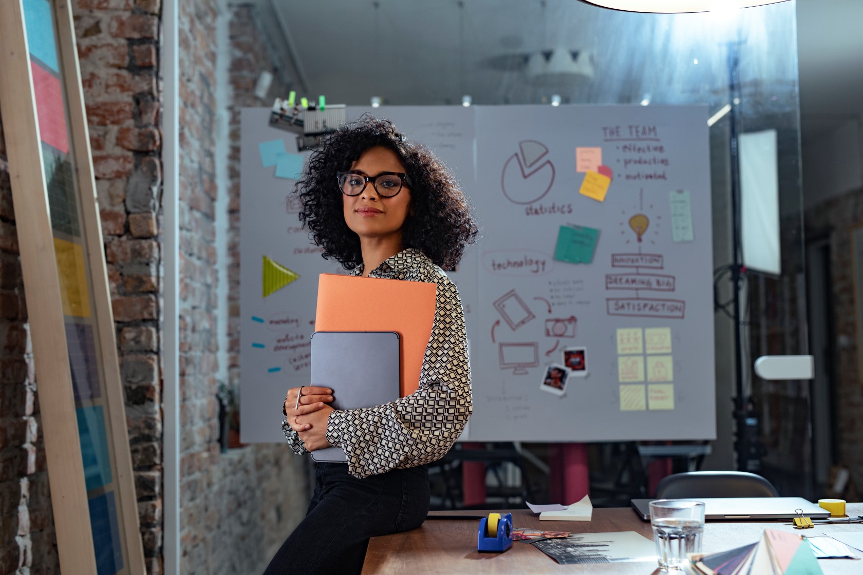 Woman in check patterned top, holding an orange and grey folder, standing in front of a whiteboard with retention strategy plans on