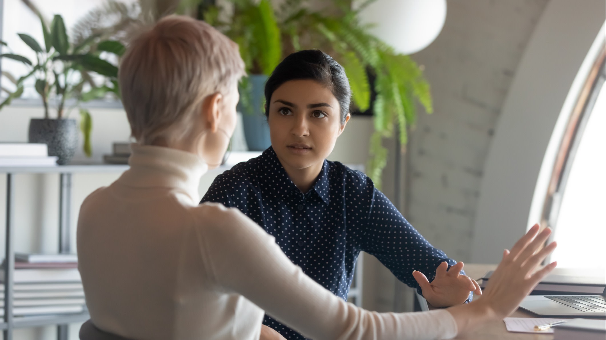 Indian and caucasian businesswomen negotiating sit at desk in office. Lawyer consulting client during formal meeting. Job interview and hiring process. Diverse colleagues discussing project concept