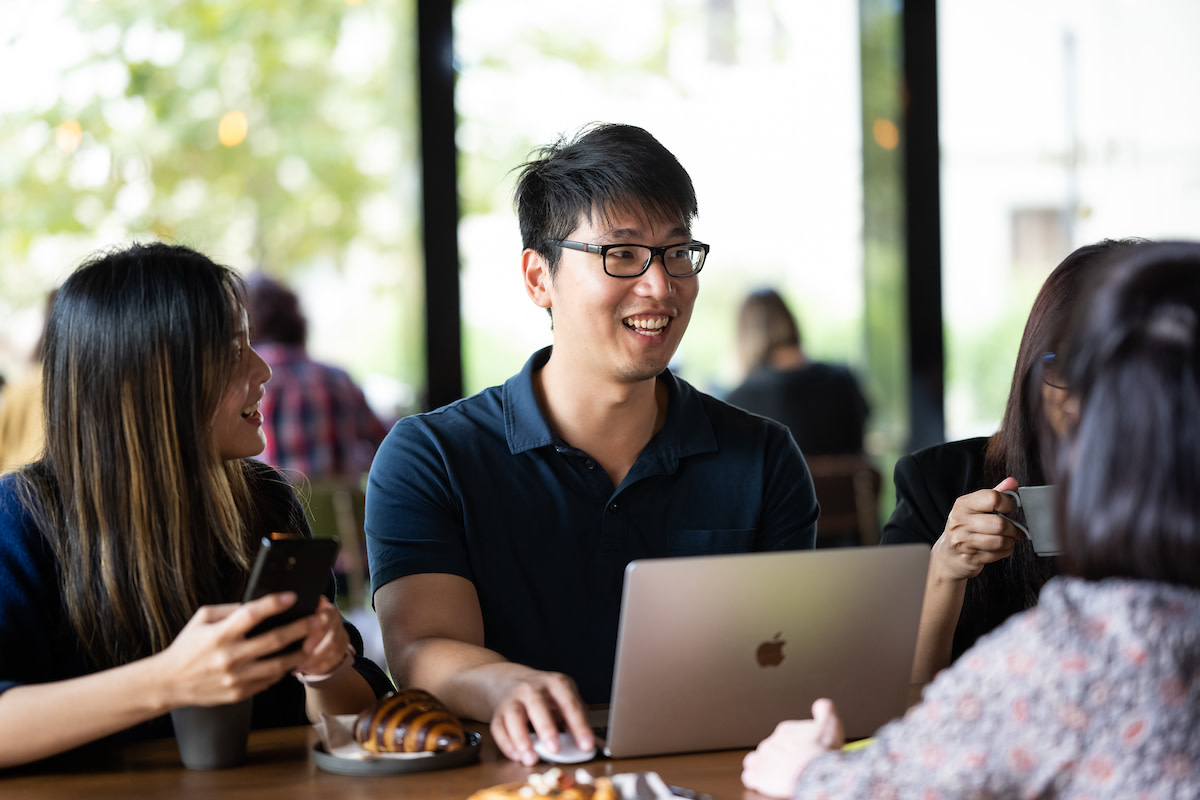 Man in a navy blue polo shirt sat smiling with his colleagues at a high table with his laptop.