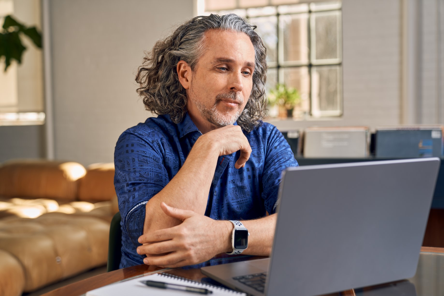 A man in a blue shirt sat with his laptop on a call with a notepad and pen beside him.