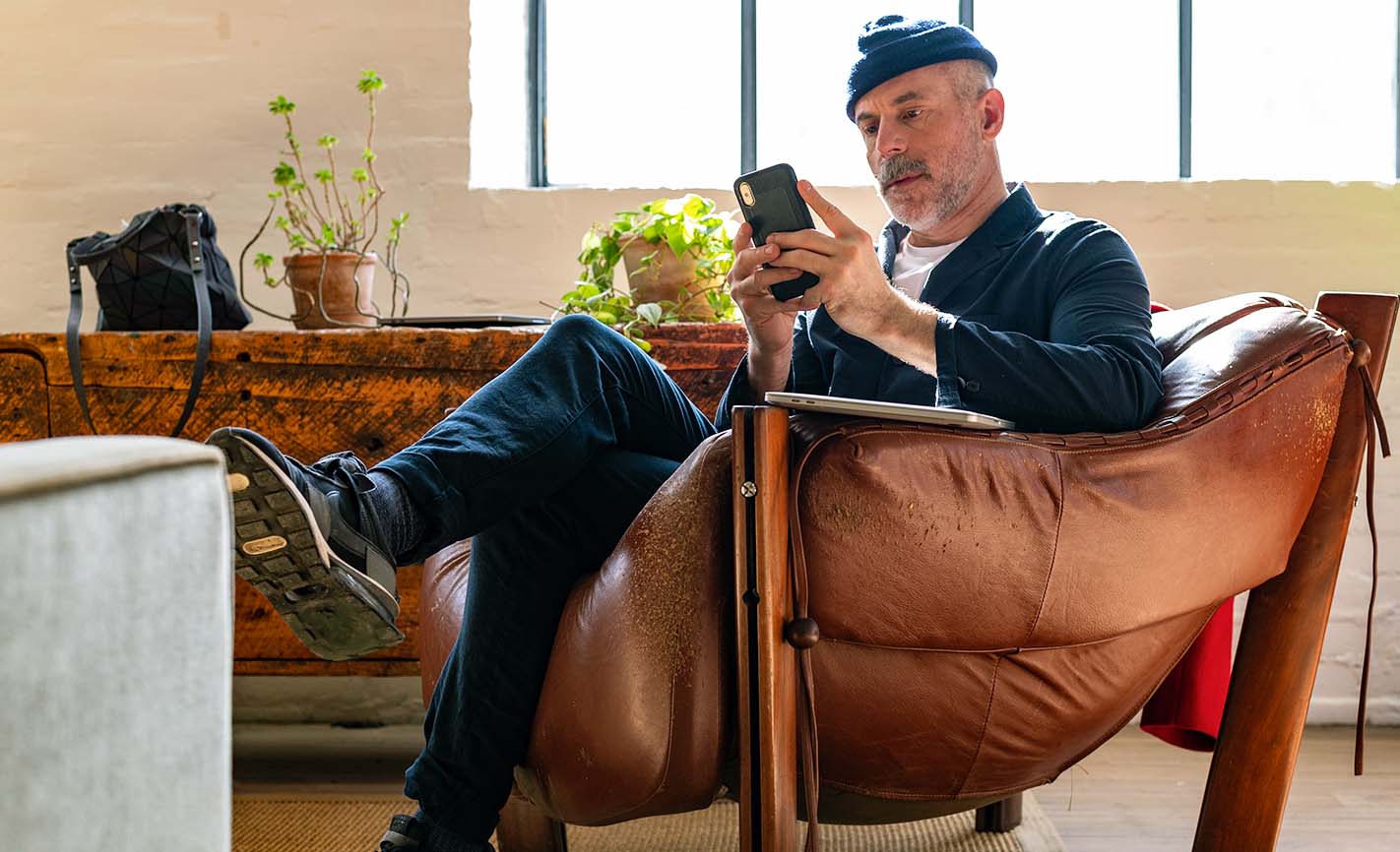 Man wearing a dark shirt over a white T-shirt, seated in a large armchair, using a digital coaching app on a mobile phone