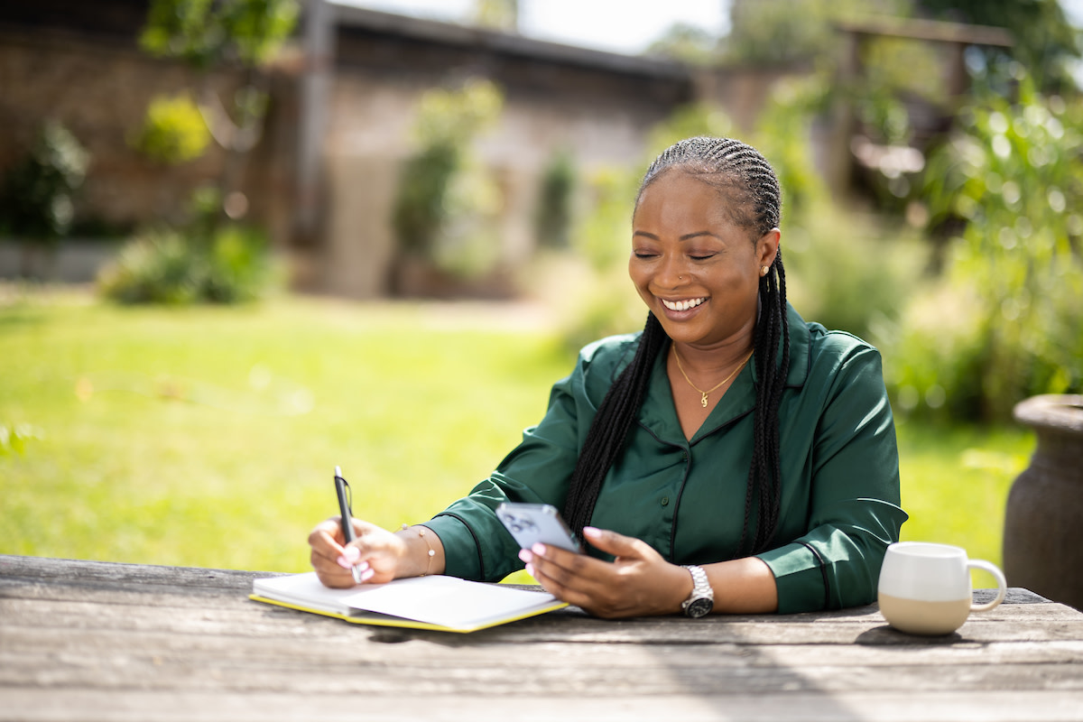 Smiling woman in a green shirt sitting outside and writing in a notebook