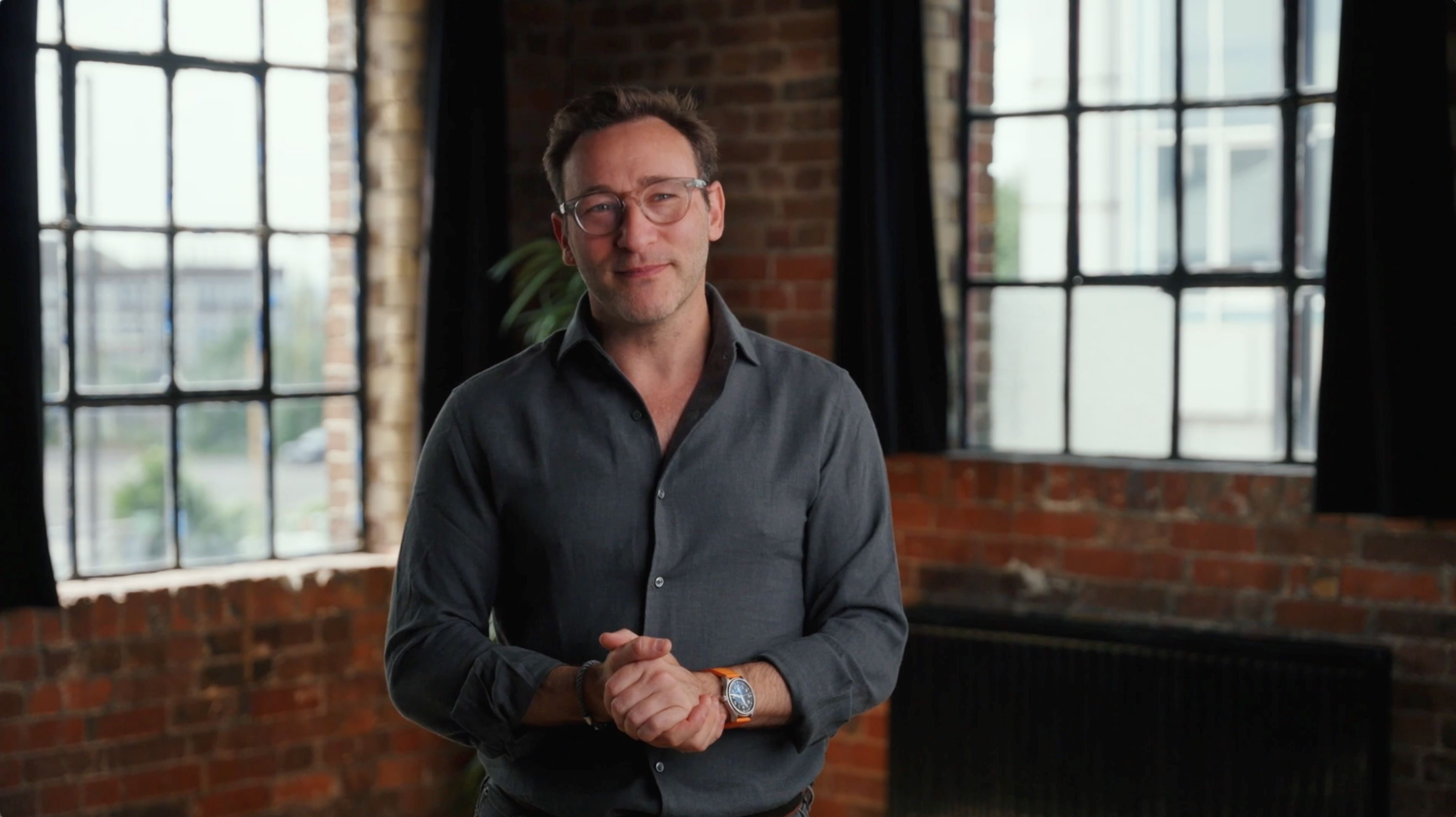 Simon Sinek wearing a grey buttoned shirt in a room with exposed brick walls and industrial windows with black curtains, smiling at the camera clasping his hands.