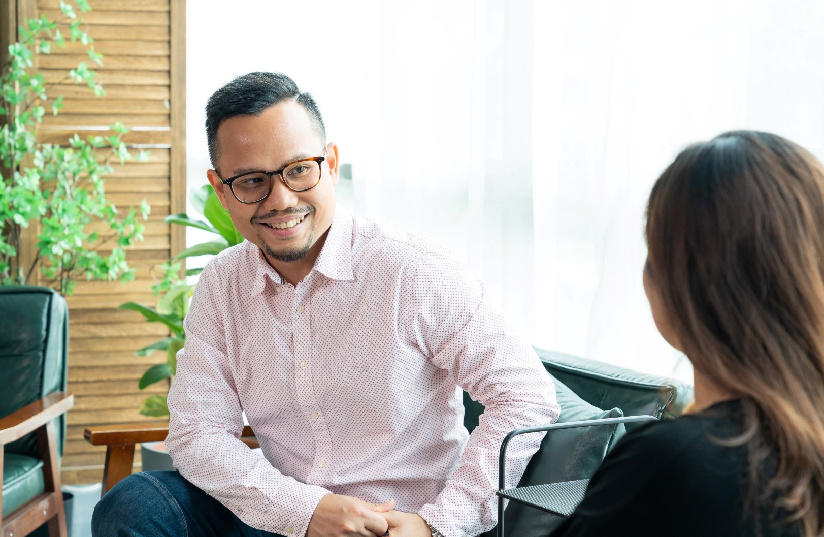 Man sat on a chair, wearing a pink shirt and glasses, facing and smiling at a colleague.