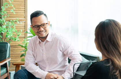 Man sat on a chair, wearing a pink shirt and glasses, facing and smiling at a colleague.